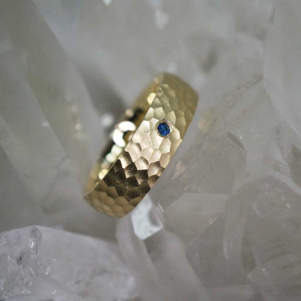 Custom made men's wedding ring in yellow gold, with a hammered texture and a single teal sapphire set into the band