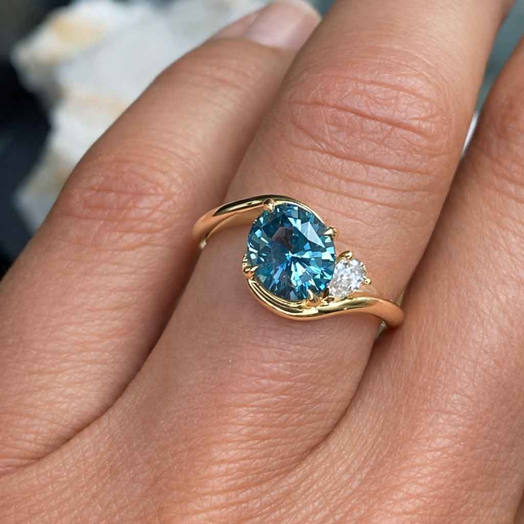 Asymmetric sapphire and diamond engagement ring in 18k yellow gold featuring an oval teal sapphire and sculptural bypass design