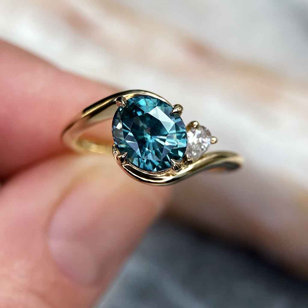 Asymmetric sapphire and diamond engagement ring in 18k yellow gold featuring an oval teal sapphire and sculptural bypass design