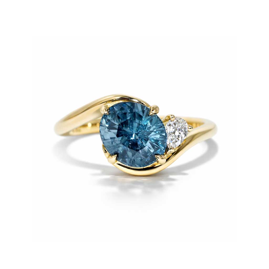 Asymmetric sapphire and diamond engagement ring in 18k yellow gold featuring an oval teal sapphire and sculptural bypass design