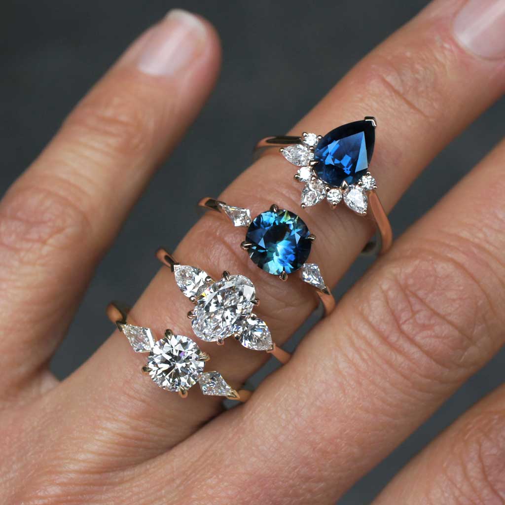 A hand stacked with precious engagement rings, featuring a round natural diamonds flanked by kite diamonds, in 18k gold trilogy design, another ring with an oval natural diamonds and a pair of pear cut diamonds, in an 18k rose gold trilogy design, the third ring features an Australian teal parti sapphire and kite diamonds in a three stone design in 18k rose gold, and the last ring is set with a pear cut teal sapphire from Madagascar and features a half halo, or a crown, of diamonds.
