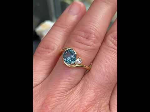 Asymmetric sapphire and diamond engagement ring in 18k yellow gold featuring an oval teal sapphire and sculptural bypass design